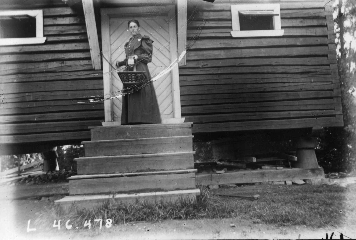 Hermann Christian Neupert, Drakt 1908-10. Kvinne stående på en trapp. I bakgrunnen et stabbur?