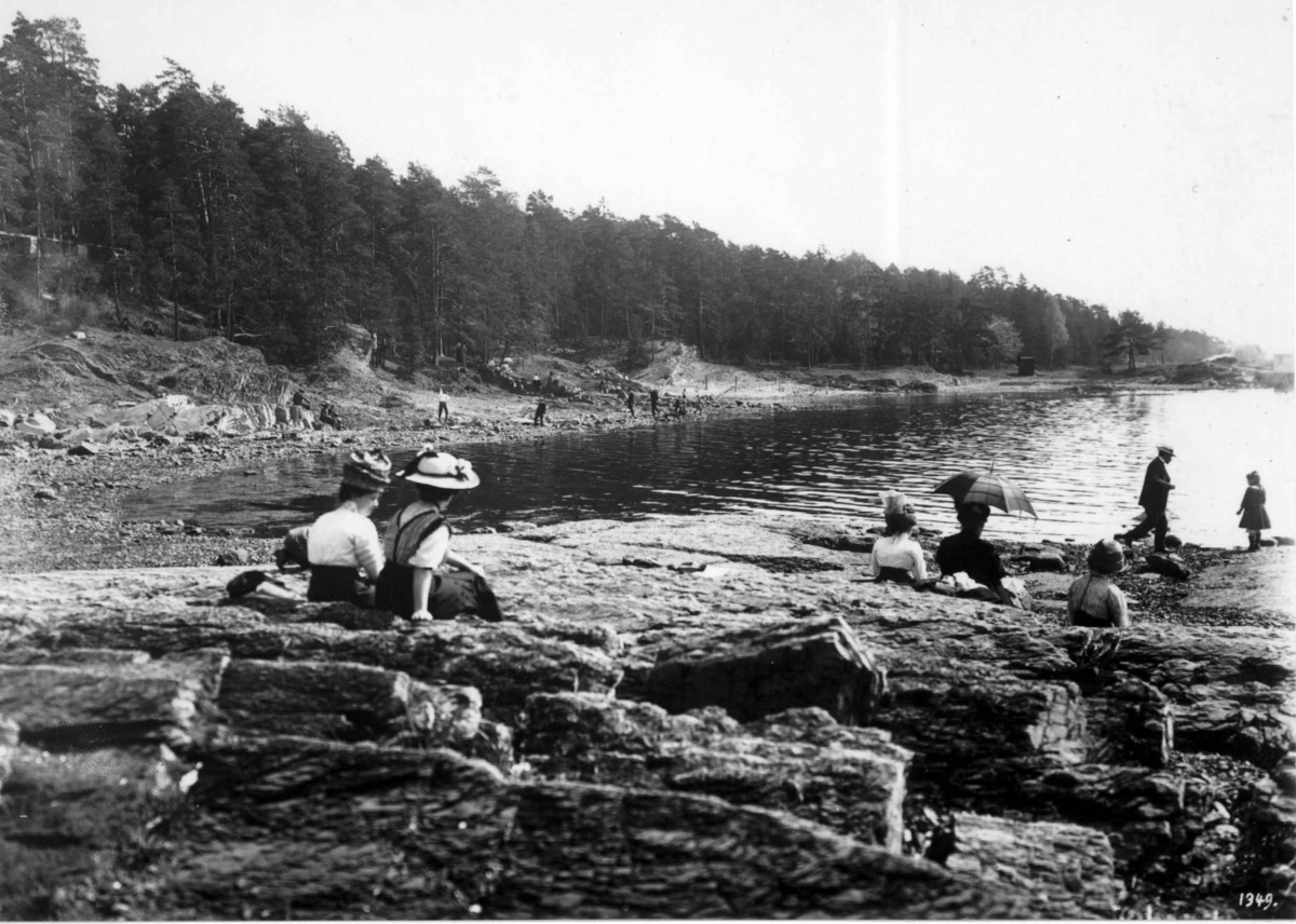Hermann Christian Neupert, Bygdøy, Oslo 1905. Sommerliv. Mennesker på stranden, antatt Paradisbukta. Mennene i dress. Kvinnene i lange kjoler eller skjørt. Alle med hatter.