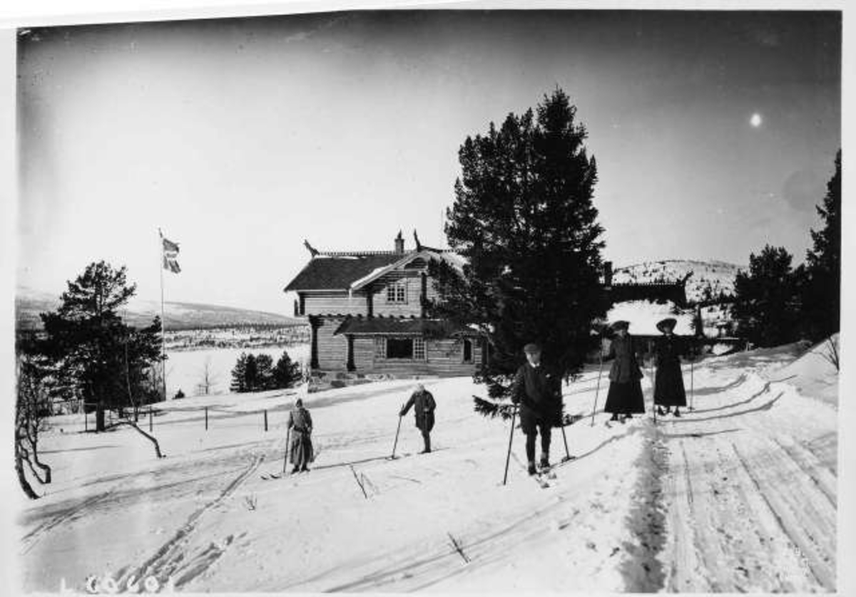Hermann Christian Neupert, Menn og kvinner på ski foran tømmerbygg i dragestil, norsk flagg vaier i vinden.
